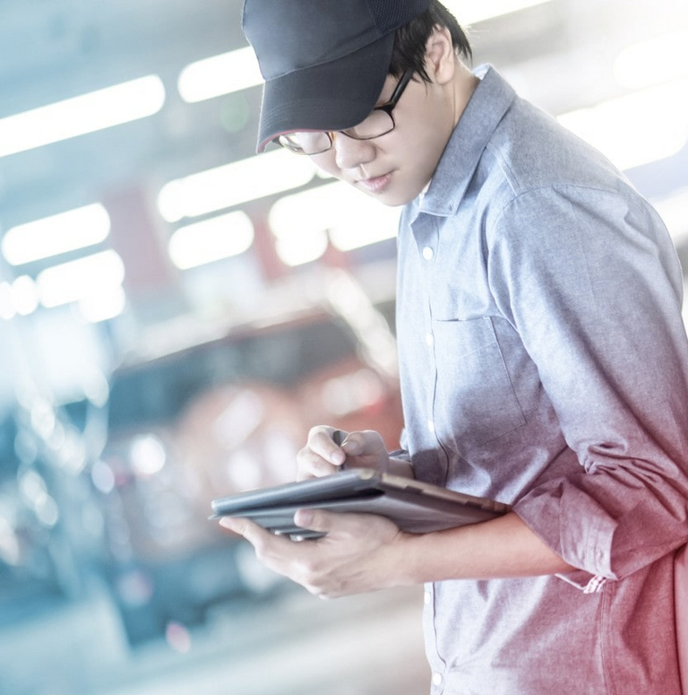 A person holding a tablet leans on a car in a factory environment.