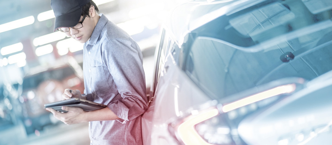 Eine Person lehnt in einer Werksumgebung an einem Auto und hält ein Tablet in der Hand.