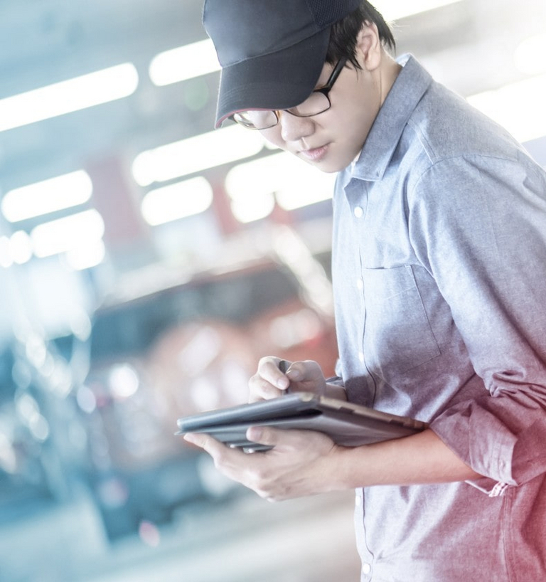 Eine Person lehnt in einer Werksumgebung an einem Auto und hält ein Tablet in der Hand.