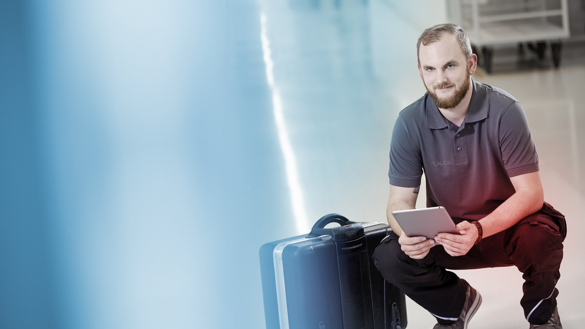 Un technicien de maintenance LAUDA est agenouillé à côté d'une valise noire et tient une tablette à la main.