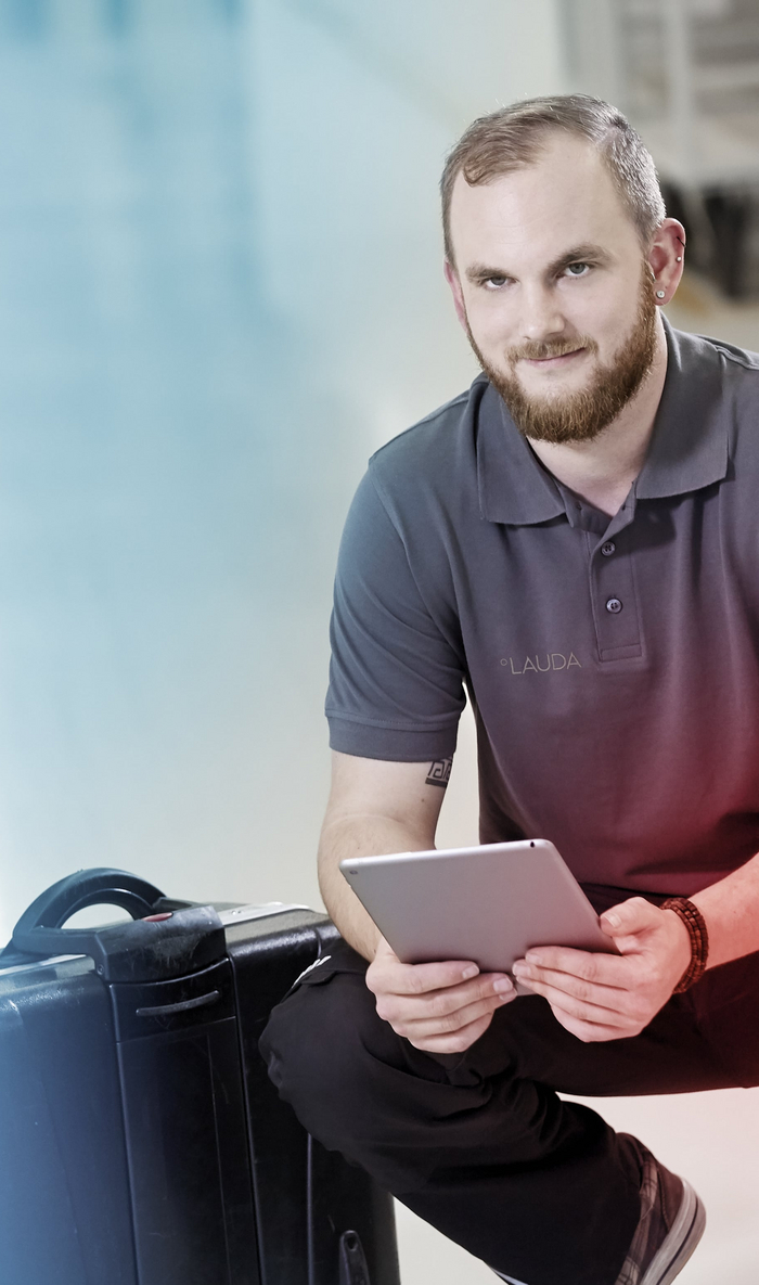 Un technicien de maintenance LAUDA est agenouillé à côté d'une valise noire et tient une tablette à la main.